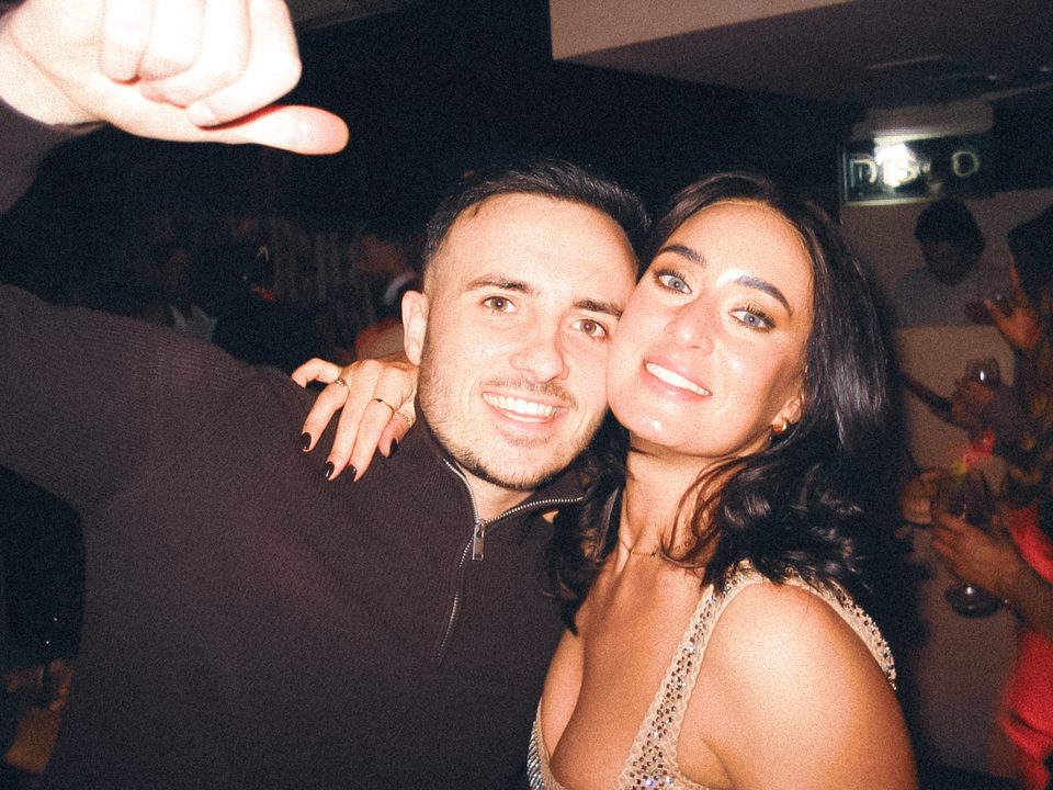 Two people posing together in a dimly lit nightclub with a crowd in background.