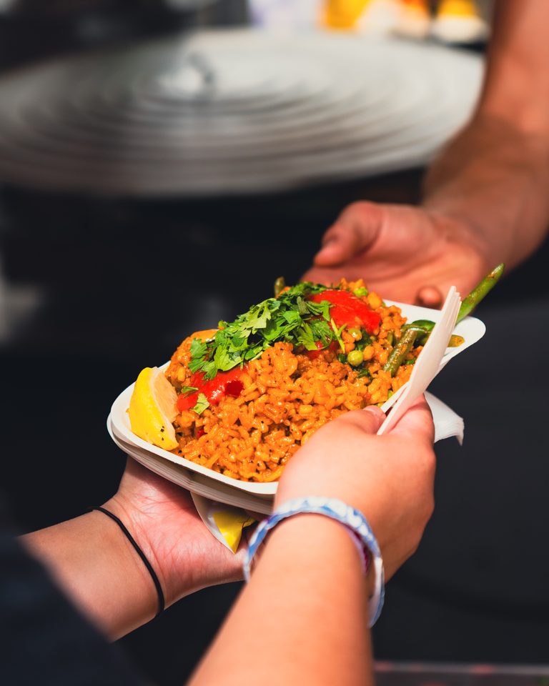 Two hands exchanging a bowl of colorful rice dish with garnishes.