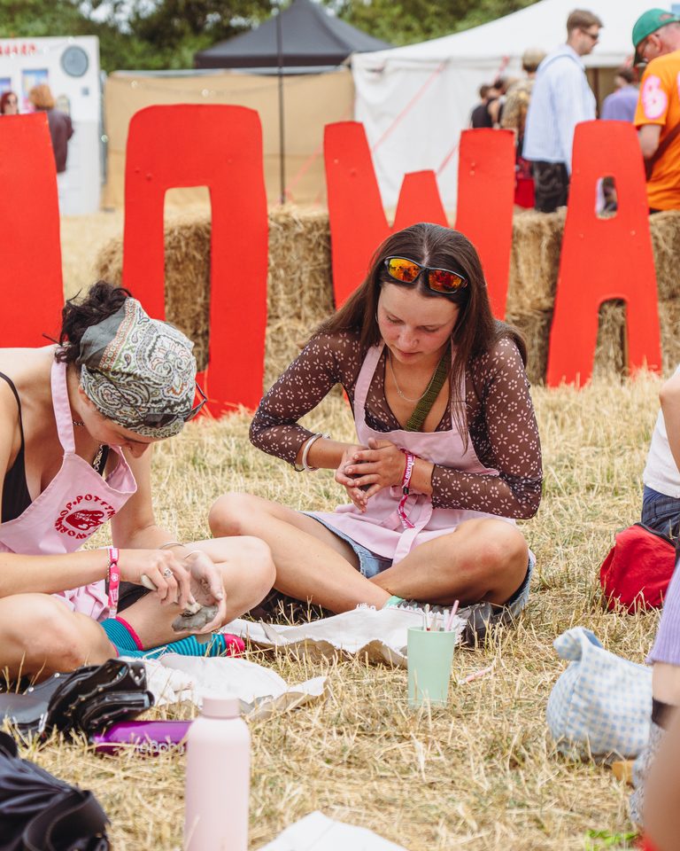 Two women sitting on grass, working with clay, with large red letters behind them.