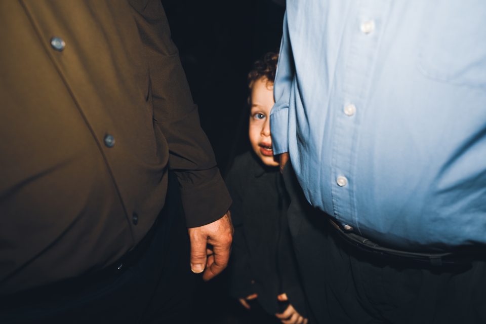 A young child peeking between two adults' legs in a dimly lit indoor space.