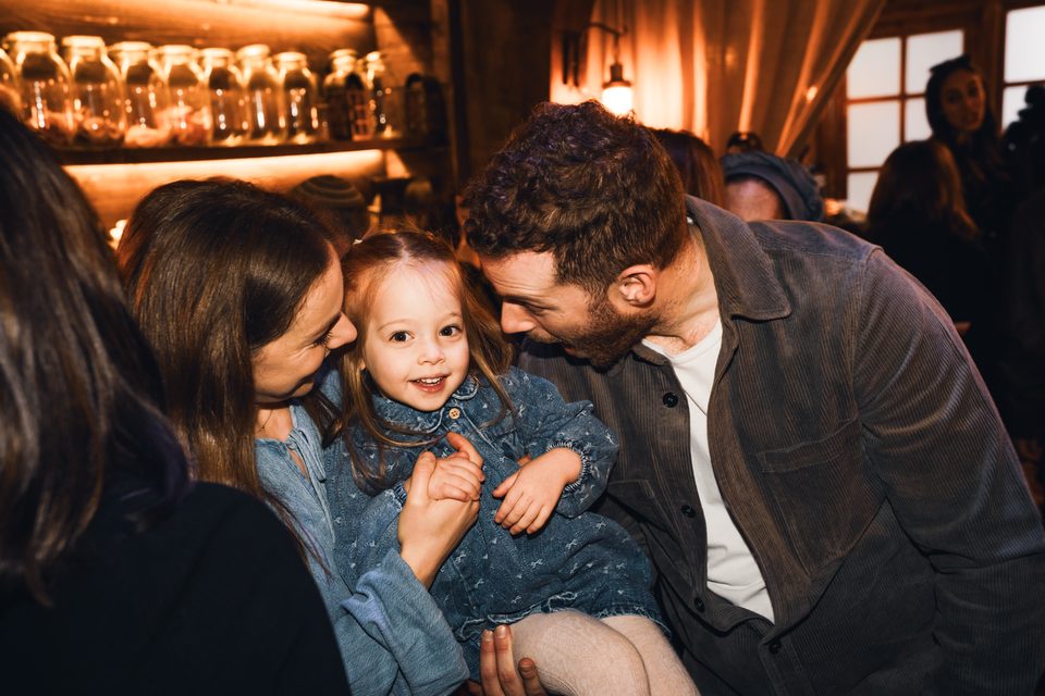 A young child held by two adults in a warmly lit indoor space with other people in background.