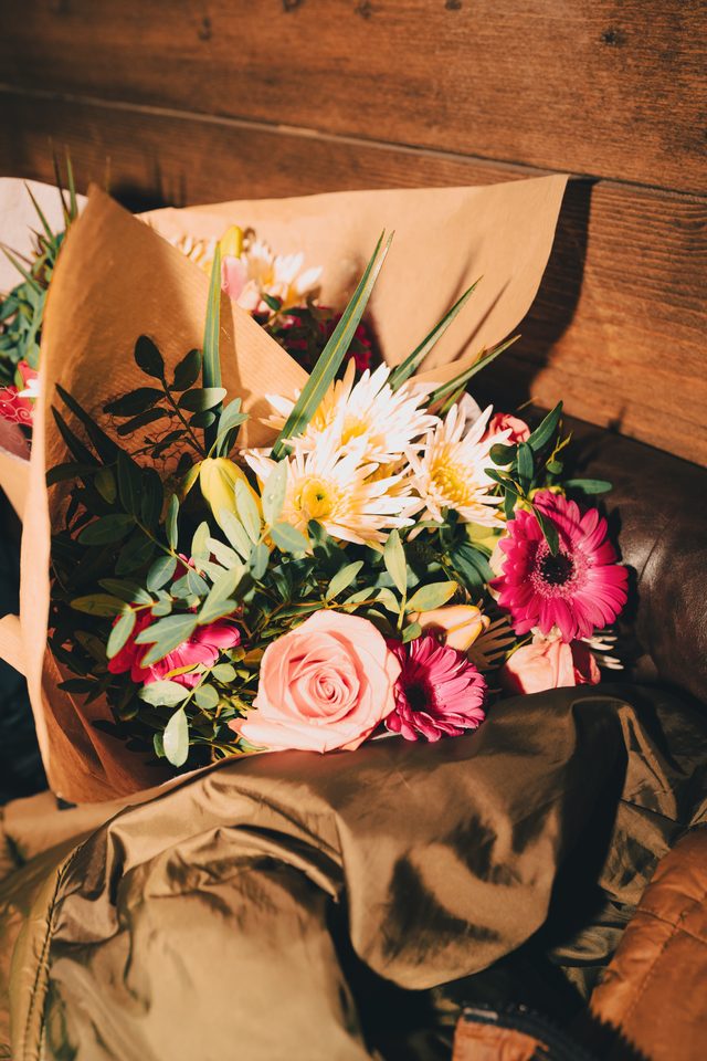 A bouquet of pink roses, white daisies, and pink gerberas wrapped in brown paper, resting on a leather surface.