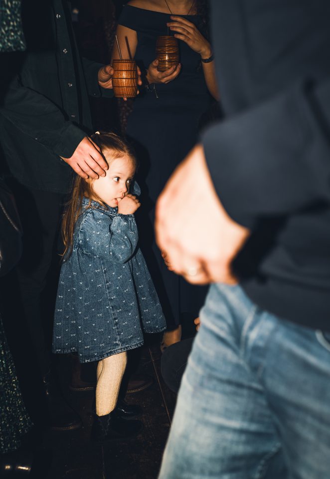 Little girl in denim dress among adults holding wooden mugs.