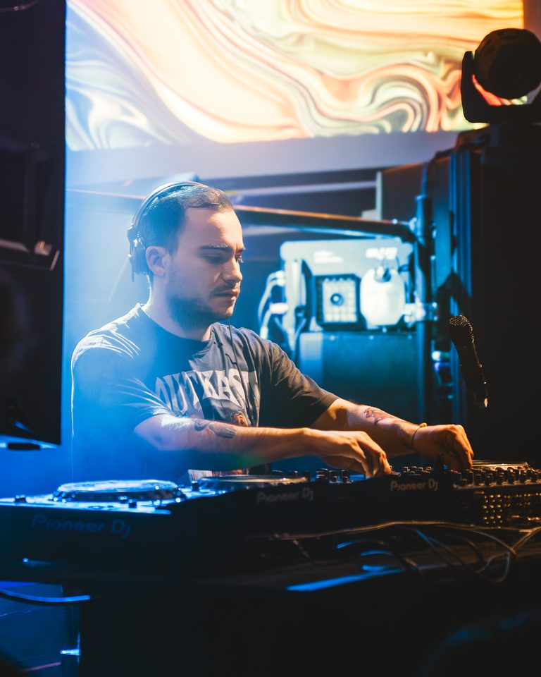 A DJ with headphones and tattoos operates Pioneer DJ equipment under blue stage lighting.