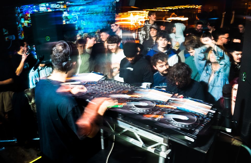A DJ with headphones at a turntable, surrounded by a crowd in a dimly lit club.