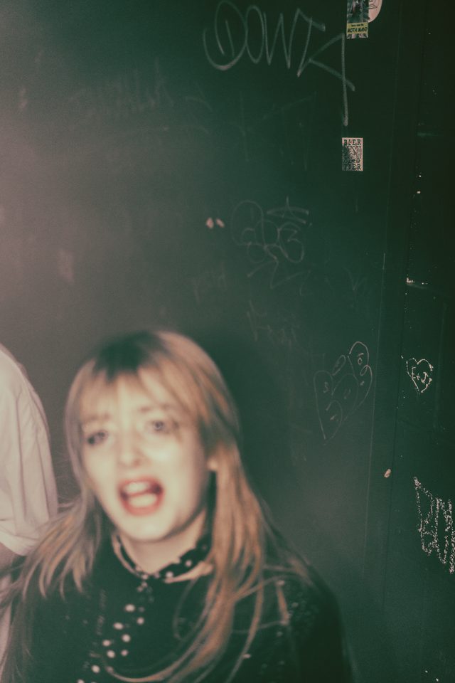 A person in a dark club, wearing a choker, with a blurred background of graffiti and stickers.