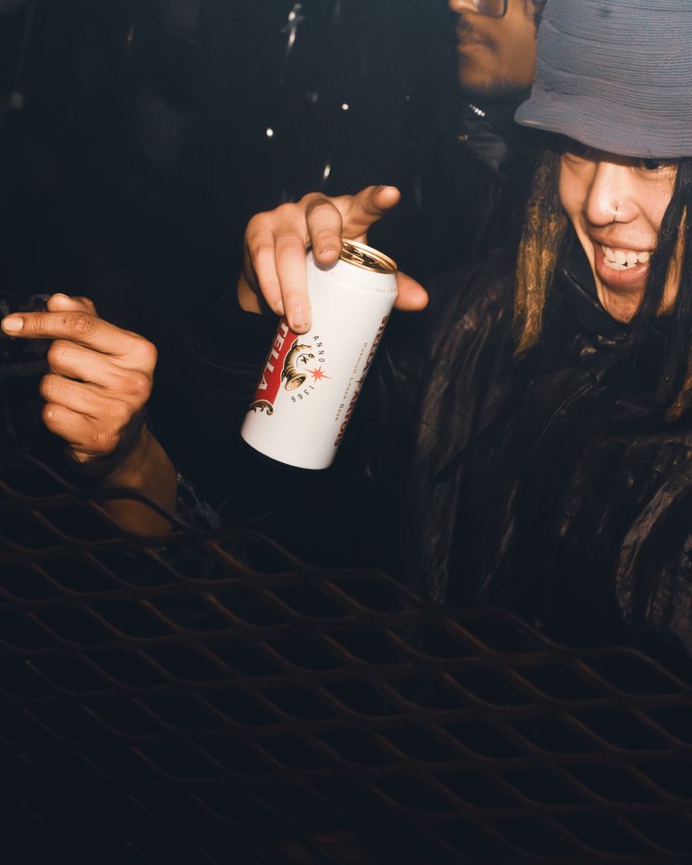 Person holding a beer can, partially obscured by a metal grate.
