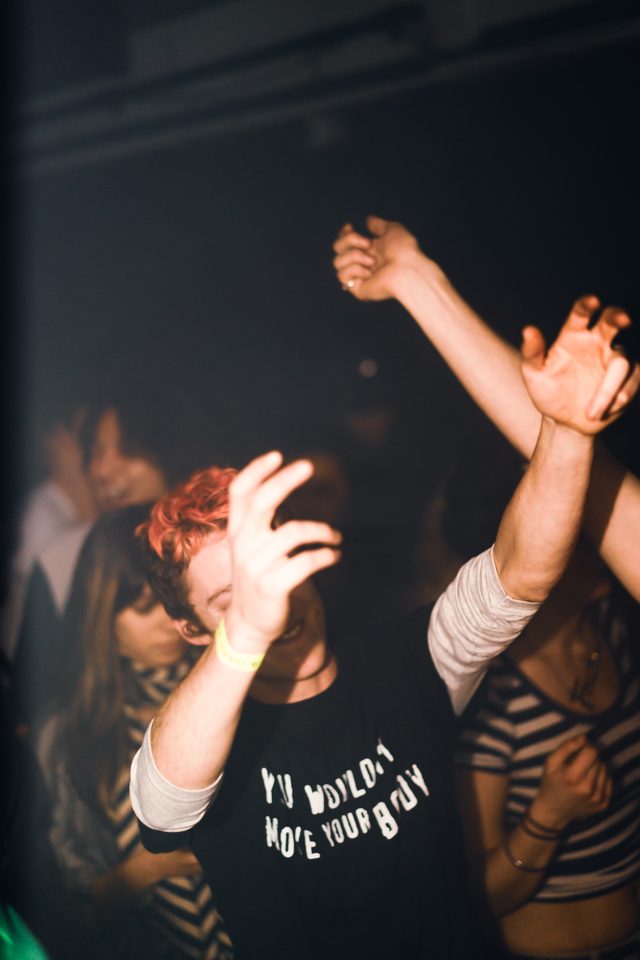 People dancing in a dark club, arms raised, one wearing a black t-shirt with text.