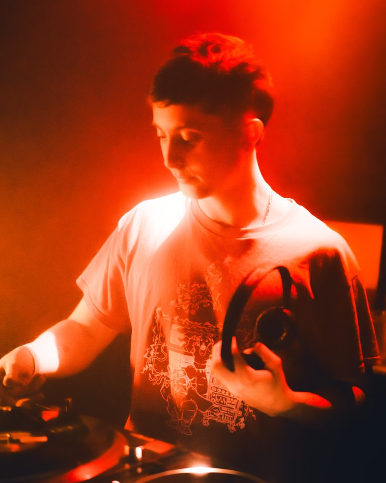 A DJ in red stage light, hands on equipment, wearing a graphic tee.