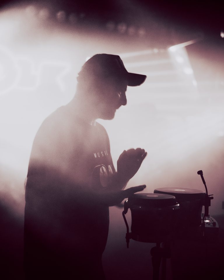 Silhouetted drummer with congas under bright stage lights.