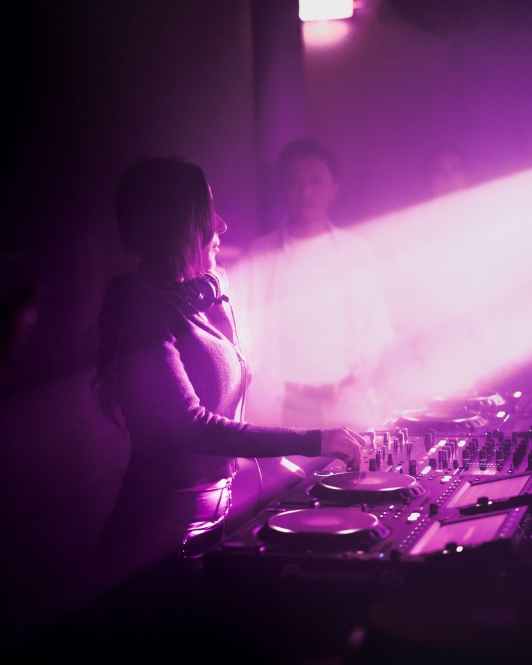 DJ behind console under purple stage light, silhouette against bright background.