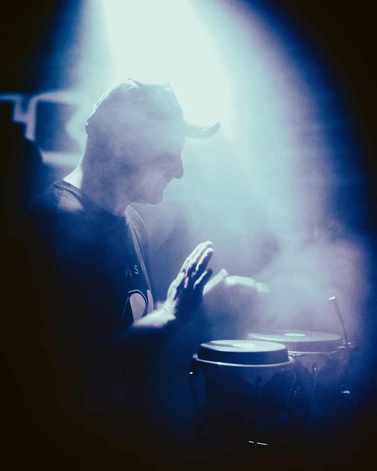 A drummer in profile, hands mid-motion, under blue stage lighting with smoke.